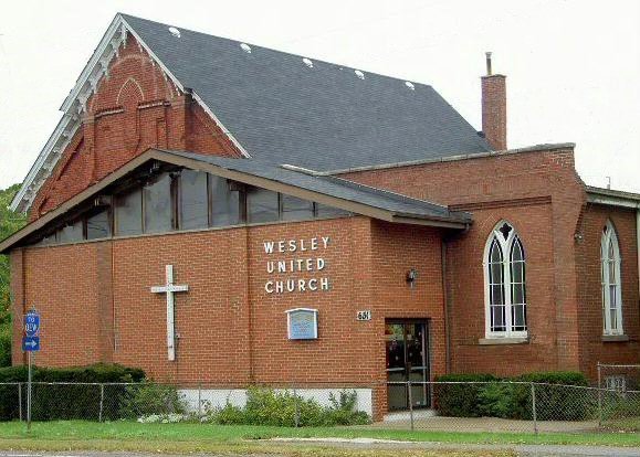 Exterior view of the brick building of Wesley United Church in Stoney Creek, showing the front entrance, a large white cross on the wall, and stained-glass windows.